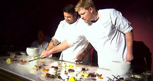 Two chefs in white uniforms preparing dishes on a large countertop in a professional kitchen, with various ingredients and tools around.