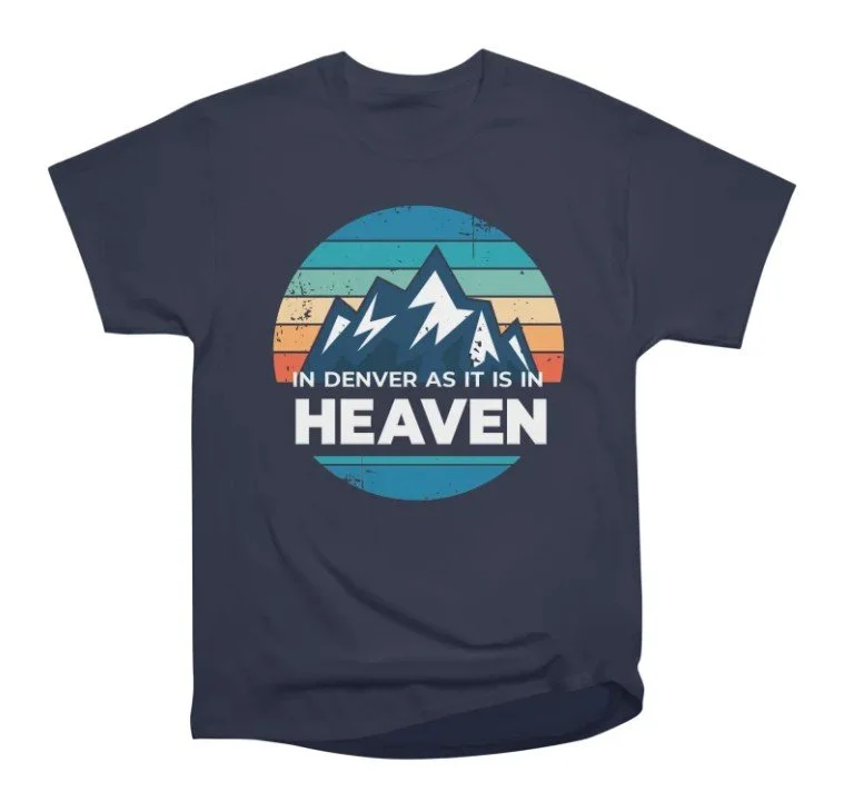 Dark blue T-shirt featuring a graphic of mountain peaks in front of a circular background with horizontal stripes in blue, green, orange, and red, and the text "In Denver As It Is In Heaven" with "Heaven" in bold.
