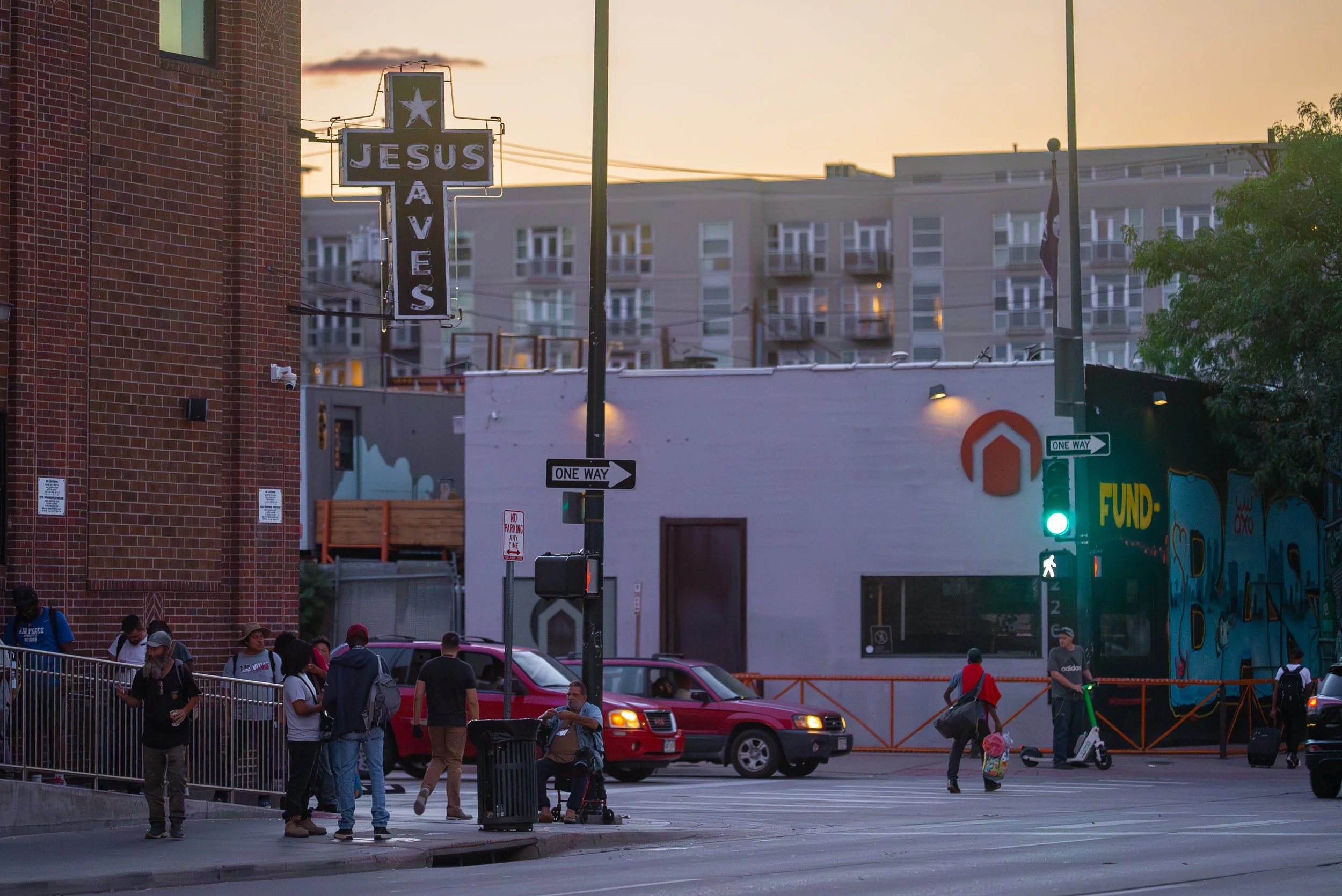 A street scene at sunset with a large sign reading 'JESUS SAVES' in the foreground. People are waiting to cross the street, cars are stopped, and a traffic light shows green for pedestrians. There is a brick building on the left, and a mural on the building in the background with bright colors and graffiti art.