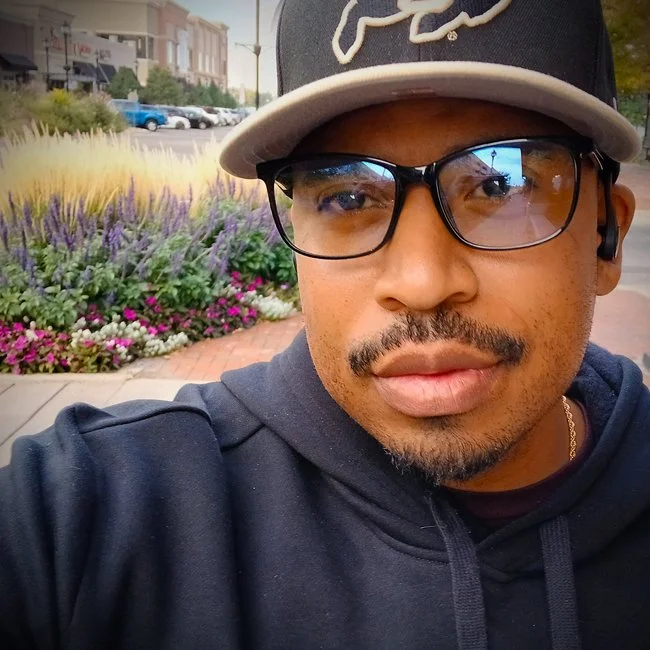 A man taking a selfie outdoors, wearing glasses, a black cap with white lettering, a dark hoodie, and earbuds. Behind him, there are colorful flowers and a city street with buildings and parked cars.