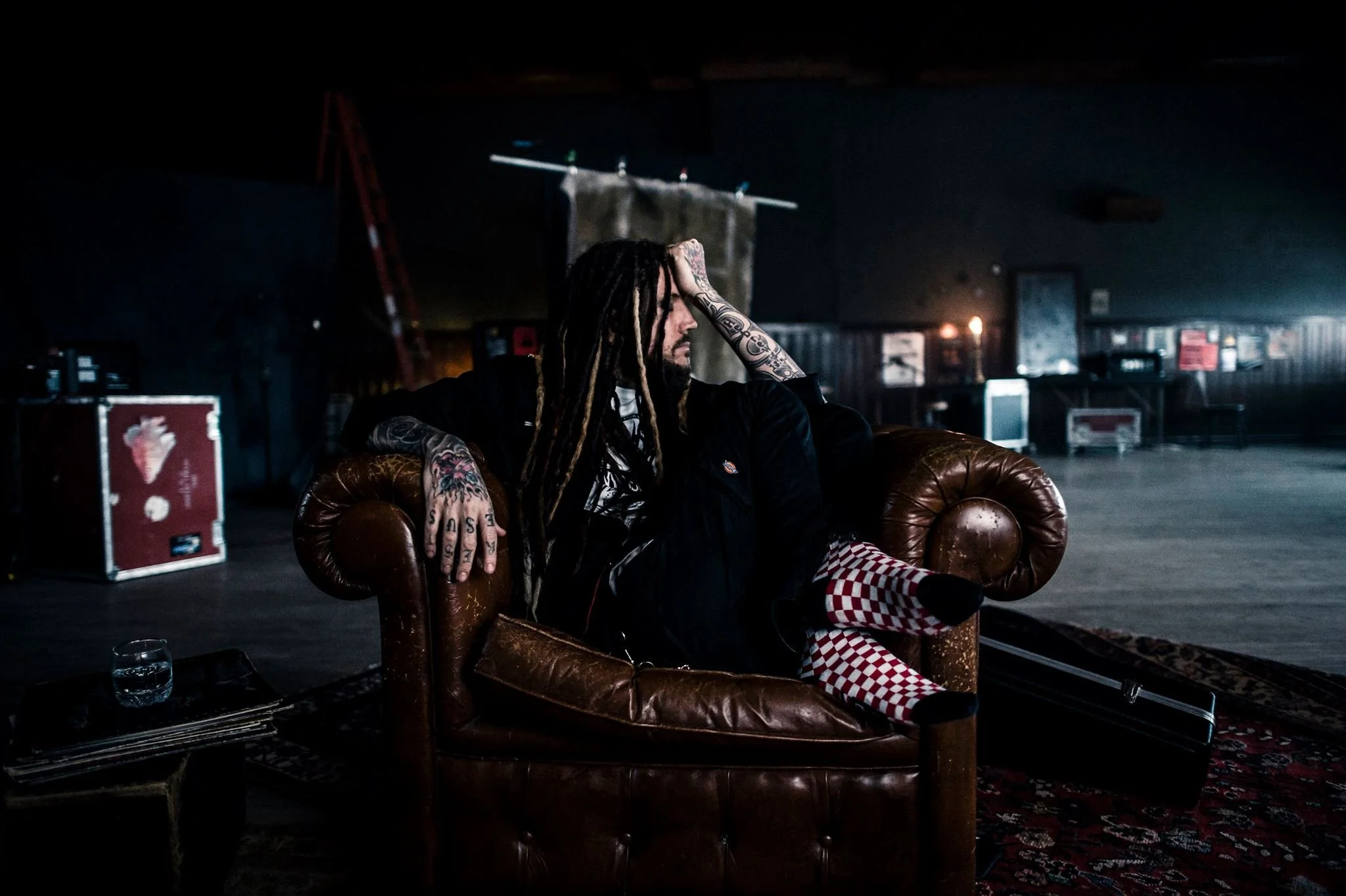 Man with long dreadlocks, tattoos, and a beard sitting in an old leather chair in a dimly lit room. He is resting his head in his hand, looking contemplative. The room has dark walls, shelves, and equipment, possibly a backstage or rehearsal space.