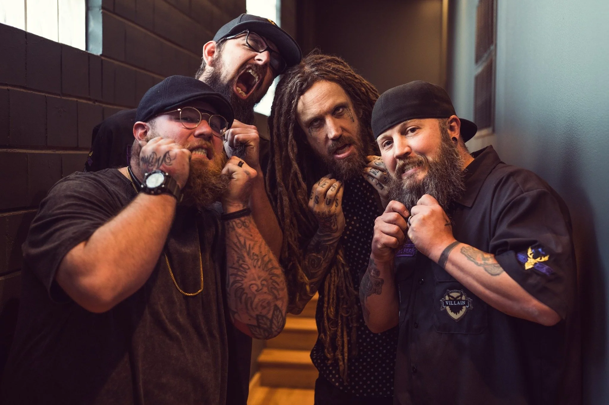 Four men with beards and tattoos posing together indoors. One man is wearing glasses and a watch, another has dreadlocks and face tattoos, the third is wearing a black hat and a vest, and the fourth is shouting or singing.