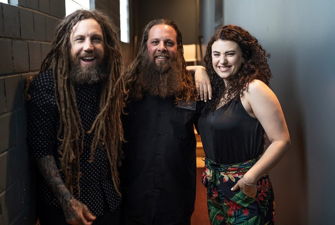 Three friends smiling and posing together in a hallway, one man with long dreadlocks and a tattoo, another man with a beard and long hair, and a woman with curly hair wearing a black top and floral pants.