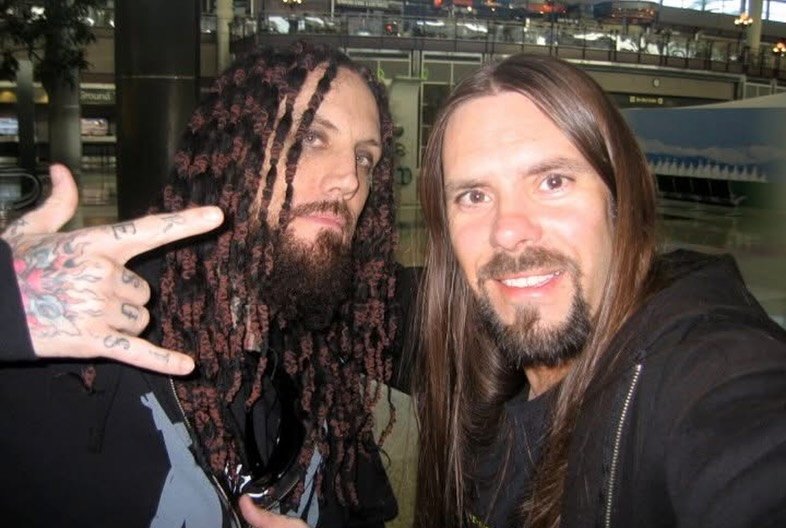 Two men posing for a selfie at an airport. One has long brown hair and a beard, the other has long dreadlocks and tattooed fingers.