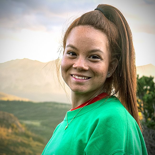 A smiling young woman with red hair tied in a high ponytail, wearing a green shirt and a necklace with a cross pendant, outdoors with mountains and trees in the background.