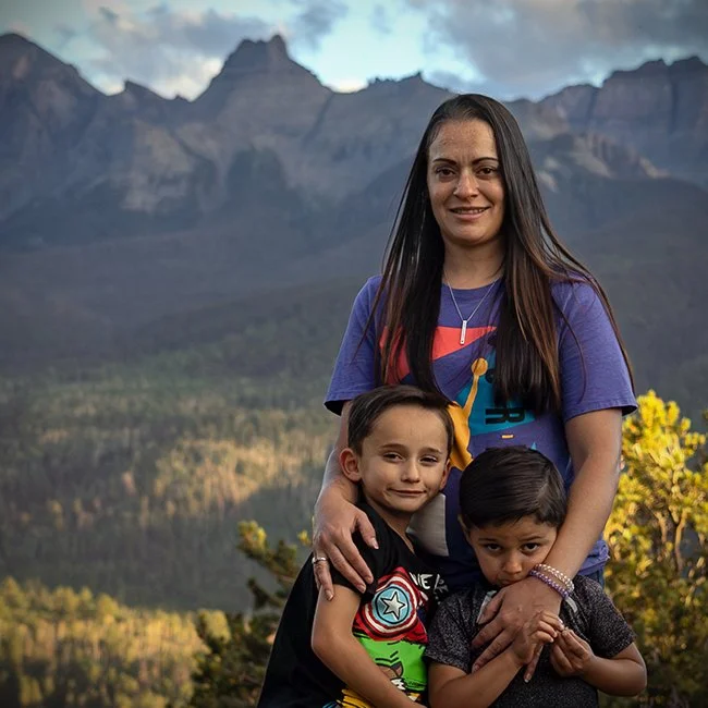 A woman with two young boys standing outdoors with a mountain range and forest in the background.