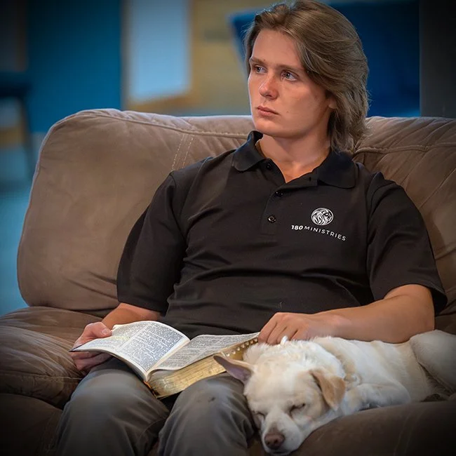 A young man with shoulder-length brown hair sitting on a beige couch with a book in his lap and a sleeping dog on his lap.