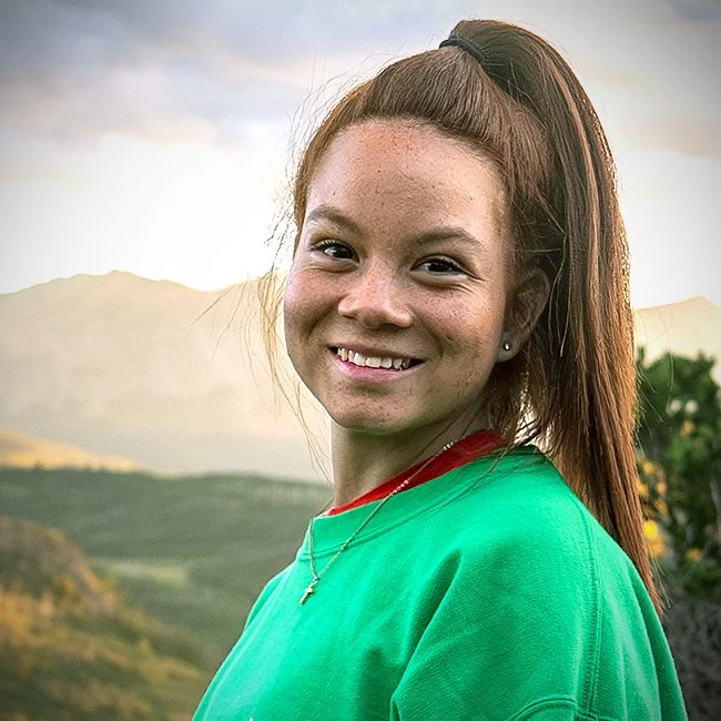 A smiling young woman with a high ponytail, wearing a green shirt and a small cross necklace, outdoors with a mountainous landscape in the background.