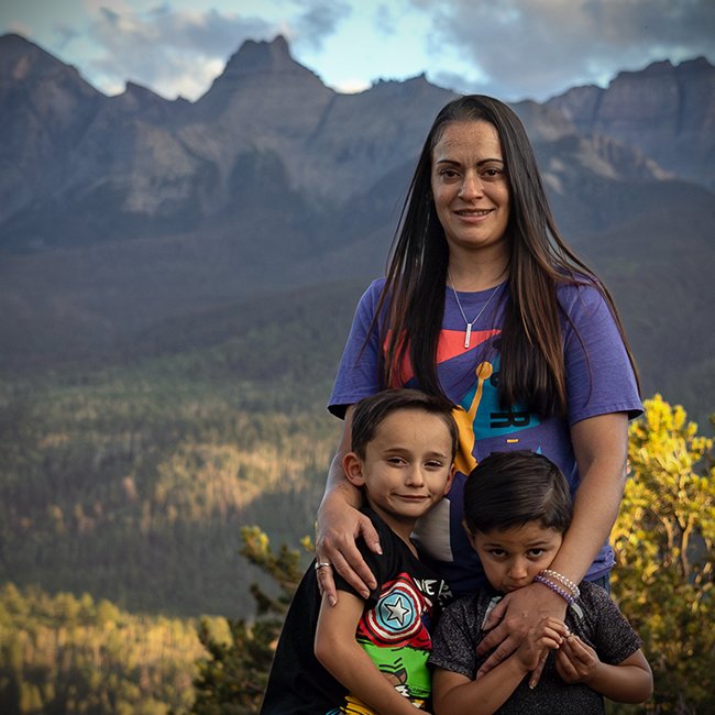 A woman with two young boys in front of a mountainous landscape with trees and a cloudy sky.