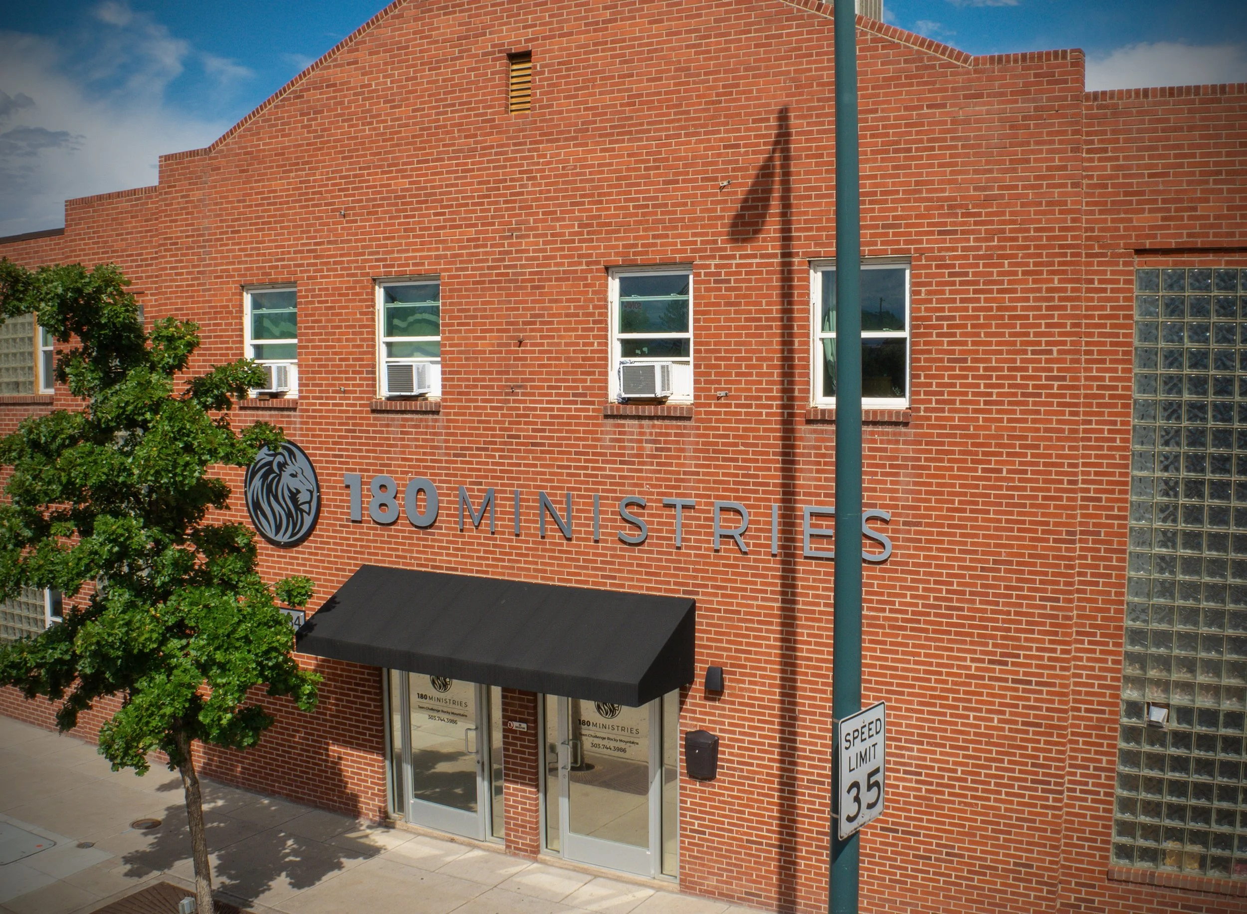 Red brick building with a sign reading '180 MINISTRIES' above the front entrance, featuring glass block windows on the right side, a lamppost casting a shadow, trees, and a speed limit sign.