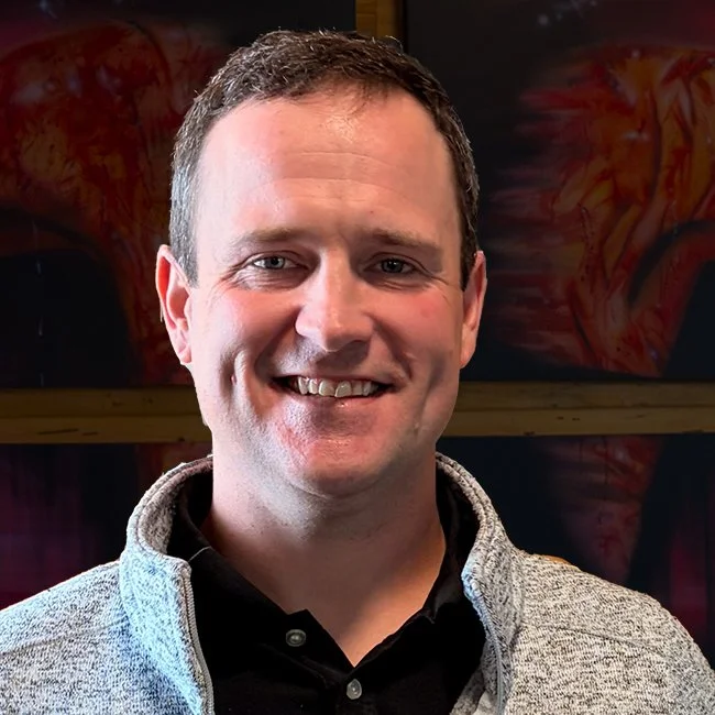 Smiling man with short dark hair, wearing a black shirt and light gray jacket, standing indoors with colorful artwork in the background.