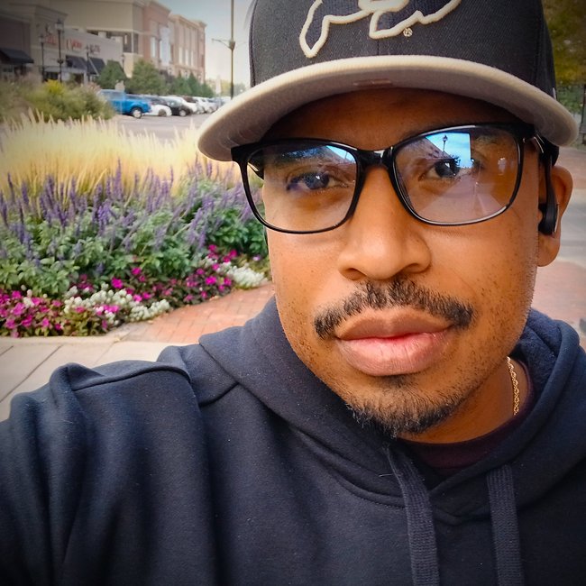 A man wearing glasses, a black baseball cap with white writing, and a dark hoodie, taking a selfie outdoors with a flower bed and shops in the background.