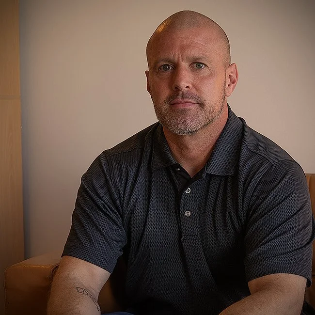 Middle-aged man with a shaved head and beard, wearing a dark polo shirt, sitting on a tan chair against a plain wall.
