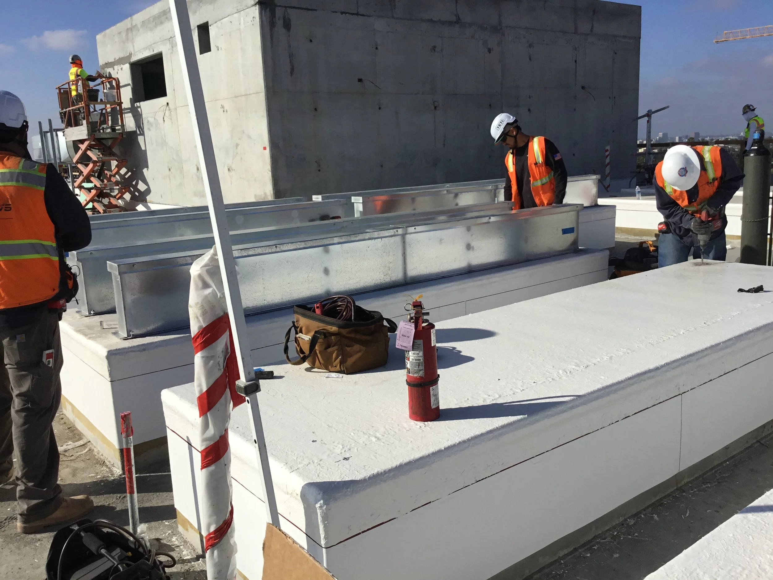 Construction workers installing or inspecting large white structural panels on a building rooftop. Workers wear safety vests and helmets, with tools and equipment nearby.