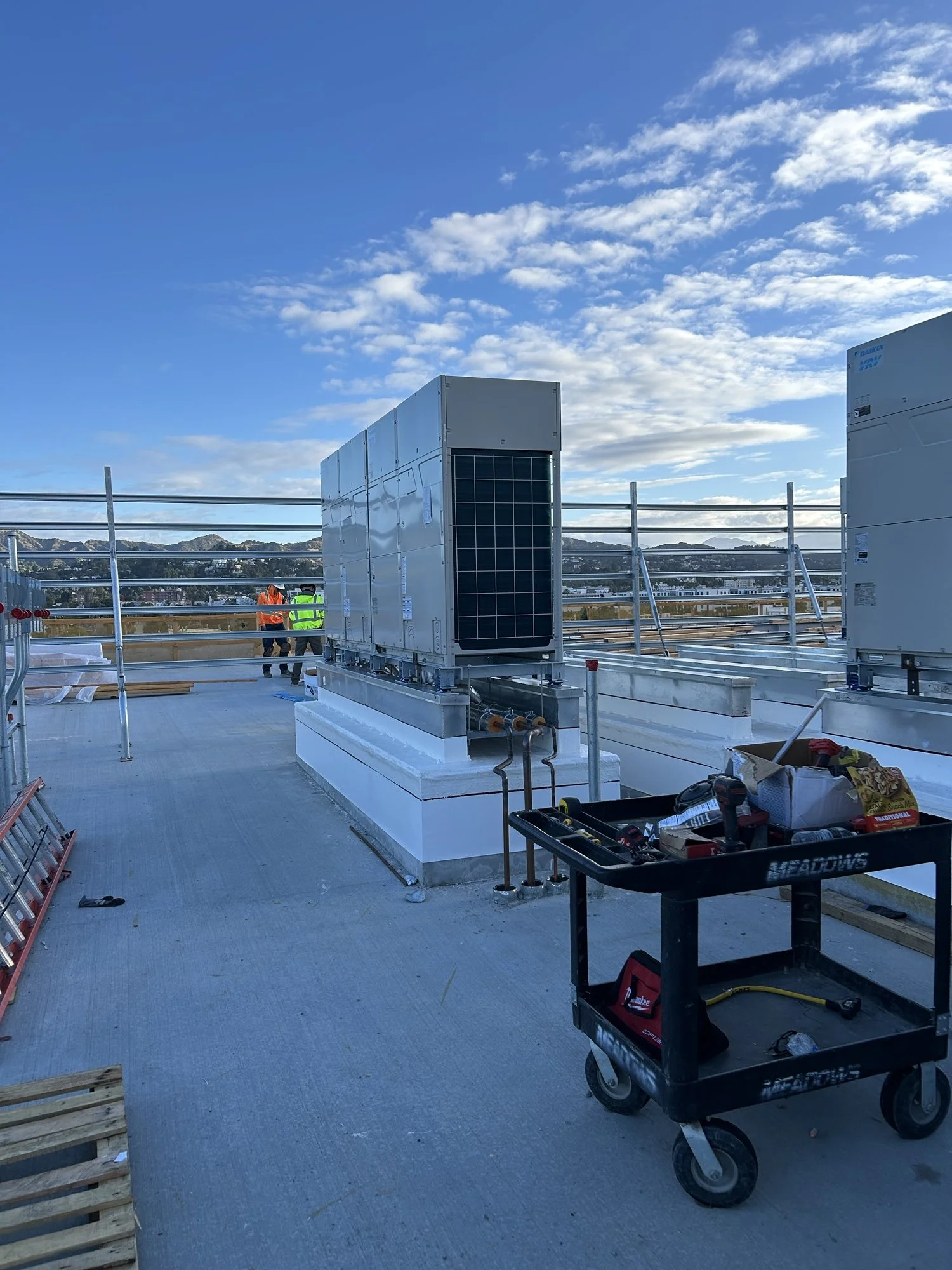 Construction workers on a rooftop installing HVAC equipment under a blue sky with clouds, with tools and supplies nearby.