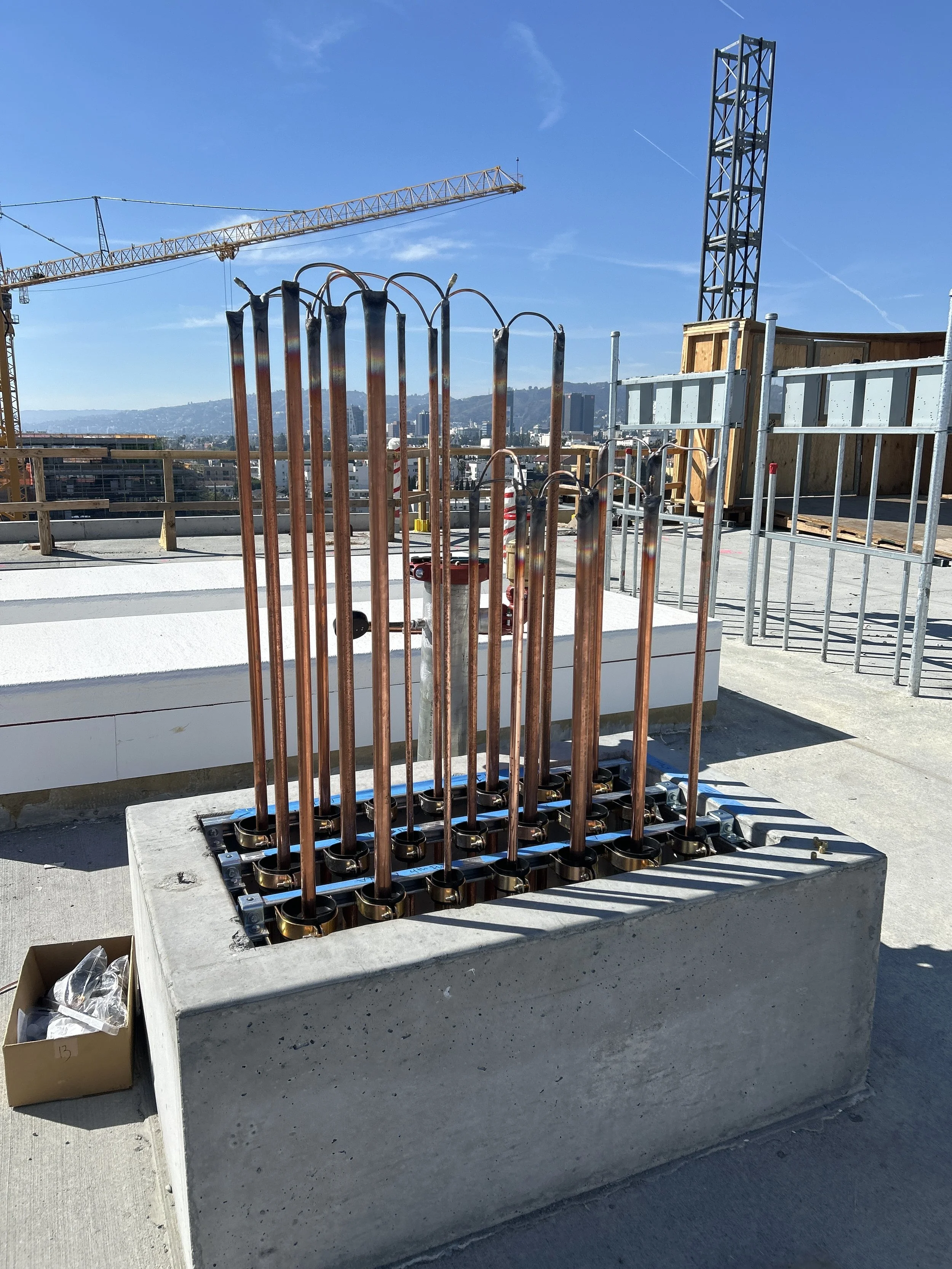 Construction site with vertical copper pipes arranged in a square base, with a crane and cityscape in the background.