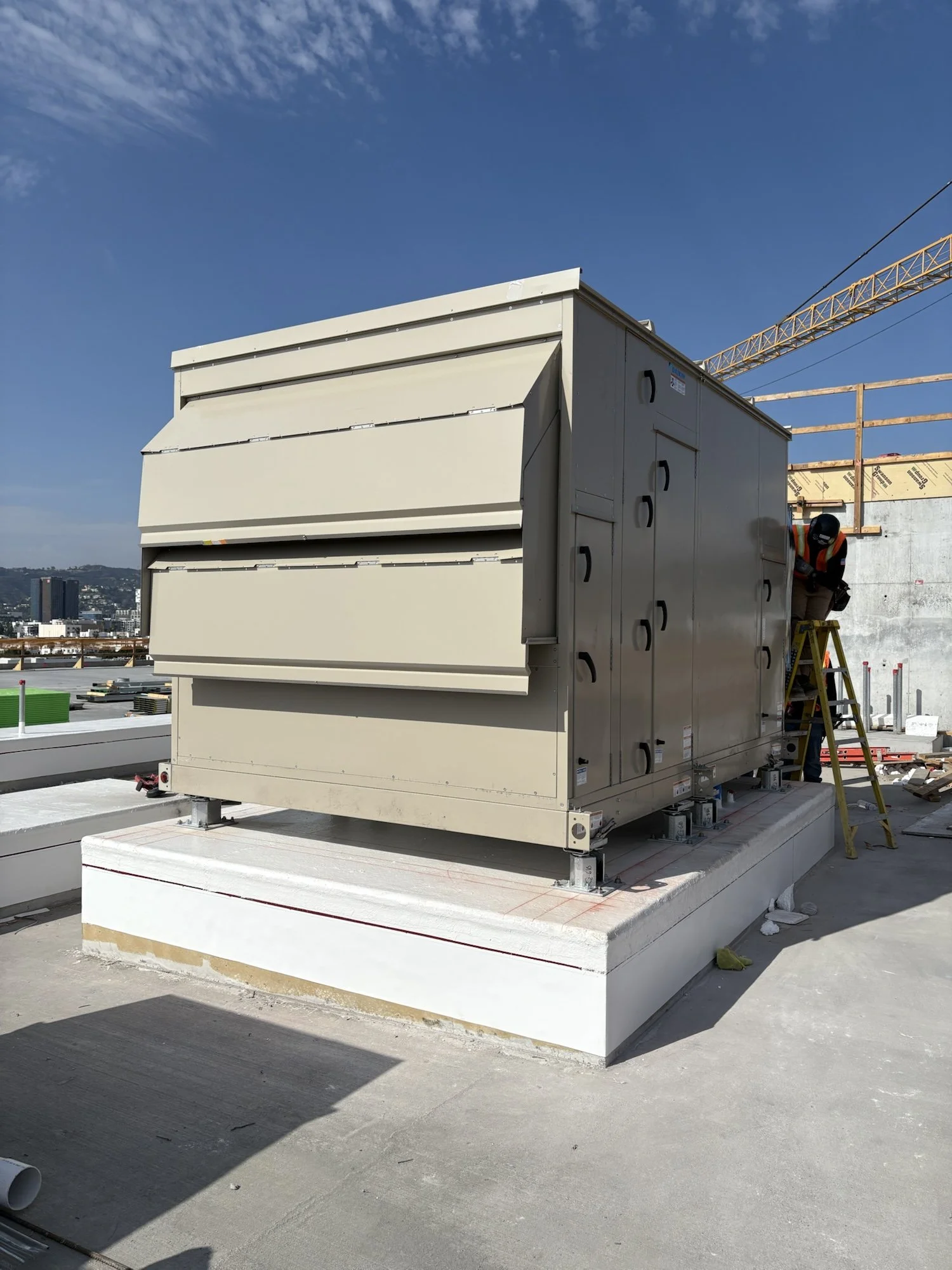 Construction worker standing on a ladder, installing or inspecting HVAC units on a building rooftop with a city skyline in the background.