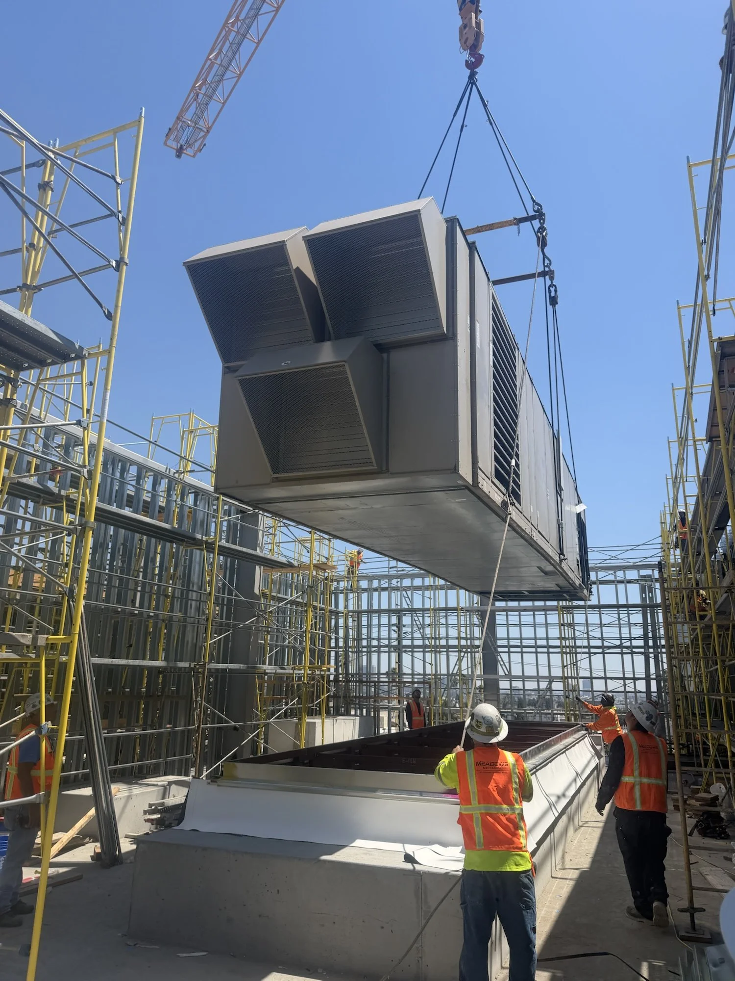 Construction workers wearing safety vests and helmets observing a large HVAC unit being lifted by a crane at a construction site with scaffolding around.