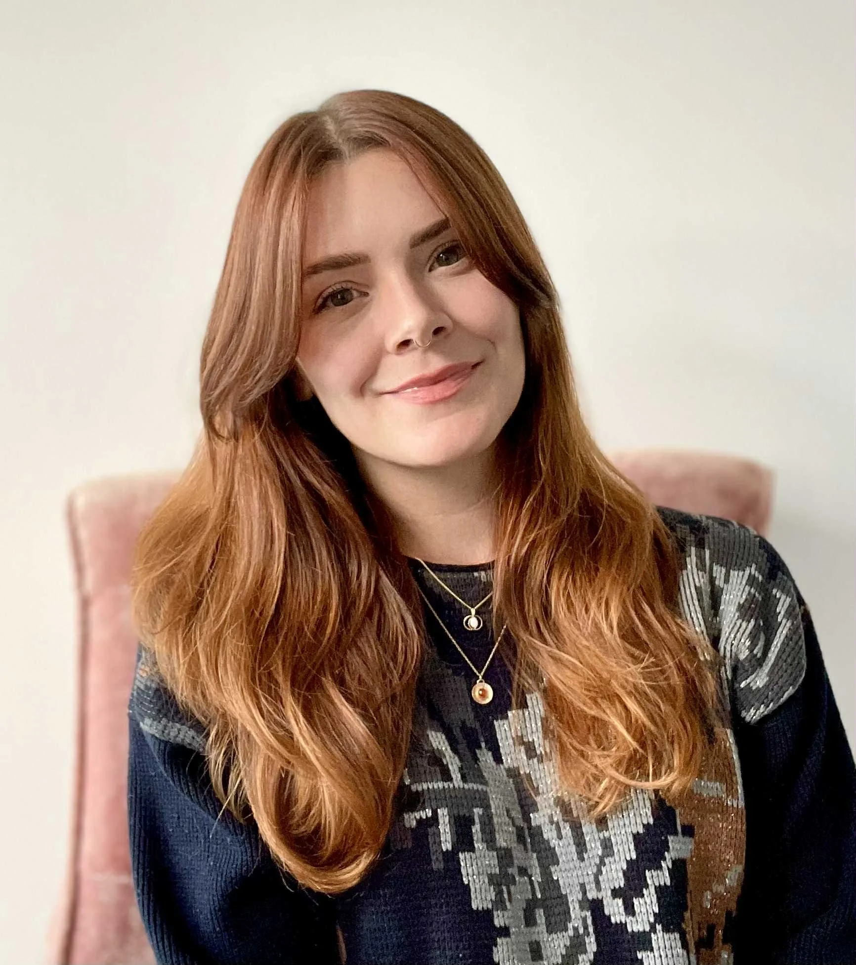 A woman with long, wavy reddish-brown hair smiling, wearing a black and white patterned top and layered gold necklaces, sitting against a light-colored wall.