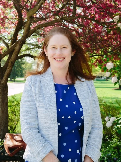 Smiling woman standing outdoors near blooming pink and white flowers, wearing a blue polka-dot dress and light gray cardigan.