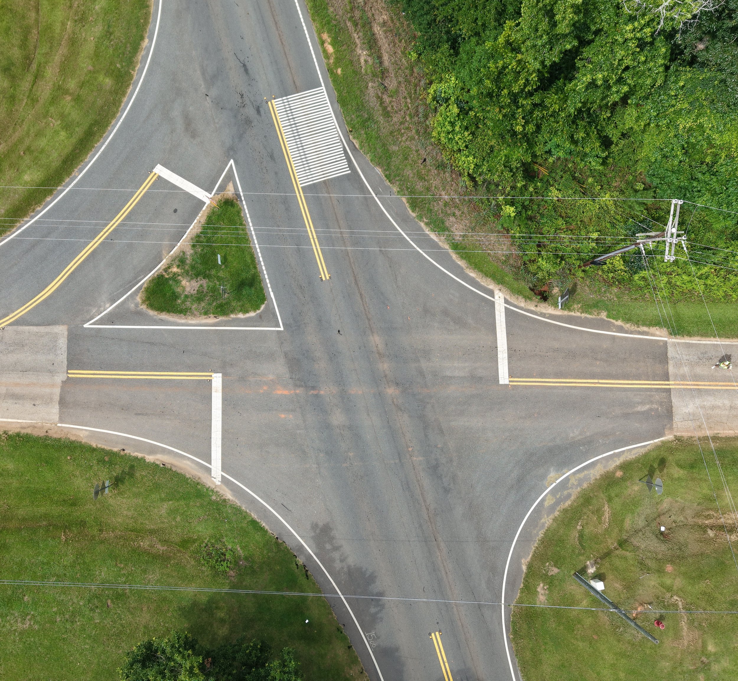 Overhead view of a curved two-lane road with double yellow lines, a pedestrian crosswalk, surrounded by green grass and trees, with utility poles and power lines crossing the scene.