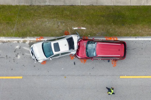An aerial view of a car accident showing a white car and a red pickup truck involved in a collision on the road. Emergency cones are placed around the scene, and a person in a reflective vest is nearby.