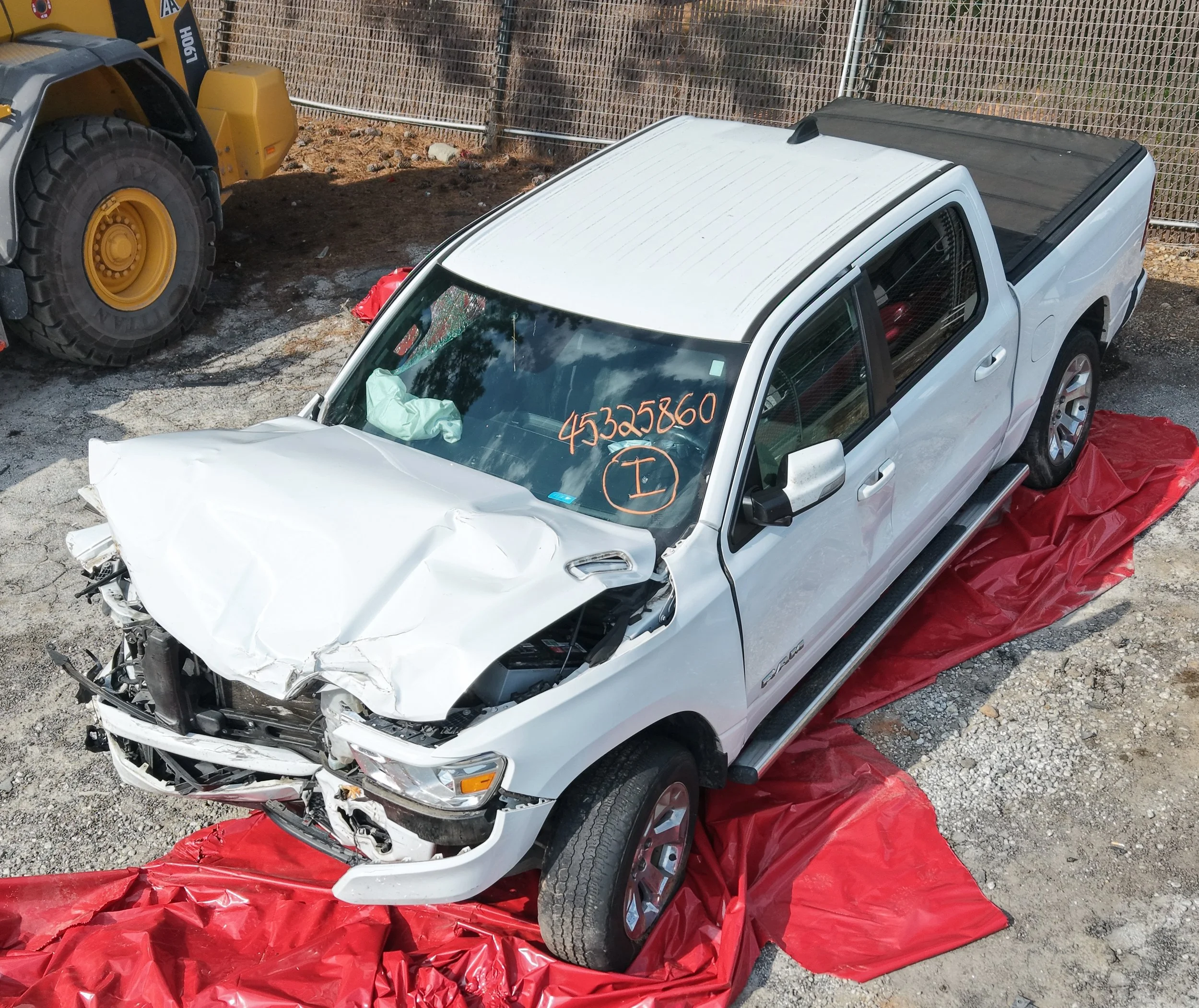 A silver pickup truck involved in a crash with significant front-end damage, parked on red crash padding on the ground, with a construction vehicle and a chain-link fence in the background.