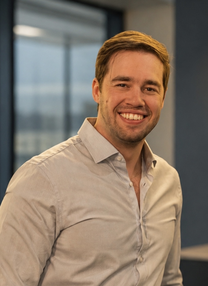A smiling man with light brown hair wearing a light beige button-up shirt standing in an office setting with a large window showing a cityscape in the background.