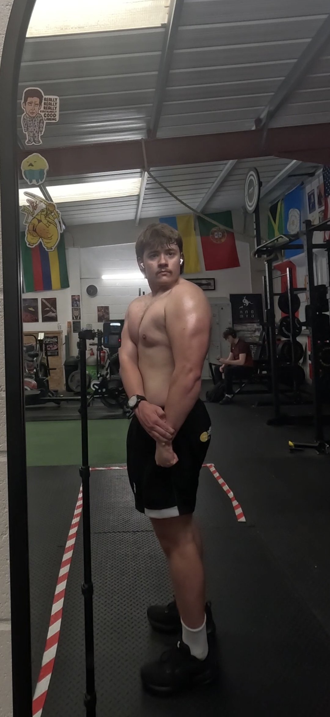 A shirtless young man with brown hair, black shorts, and white socks stands in front of a mirror in a gym. The gym has flags on the wall, exercise equipment, and a woman sitting on a bench with earphones.