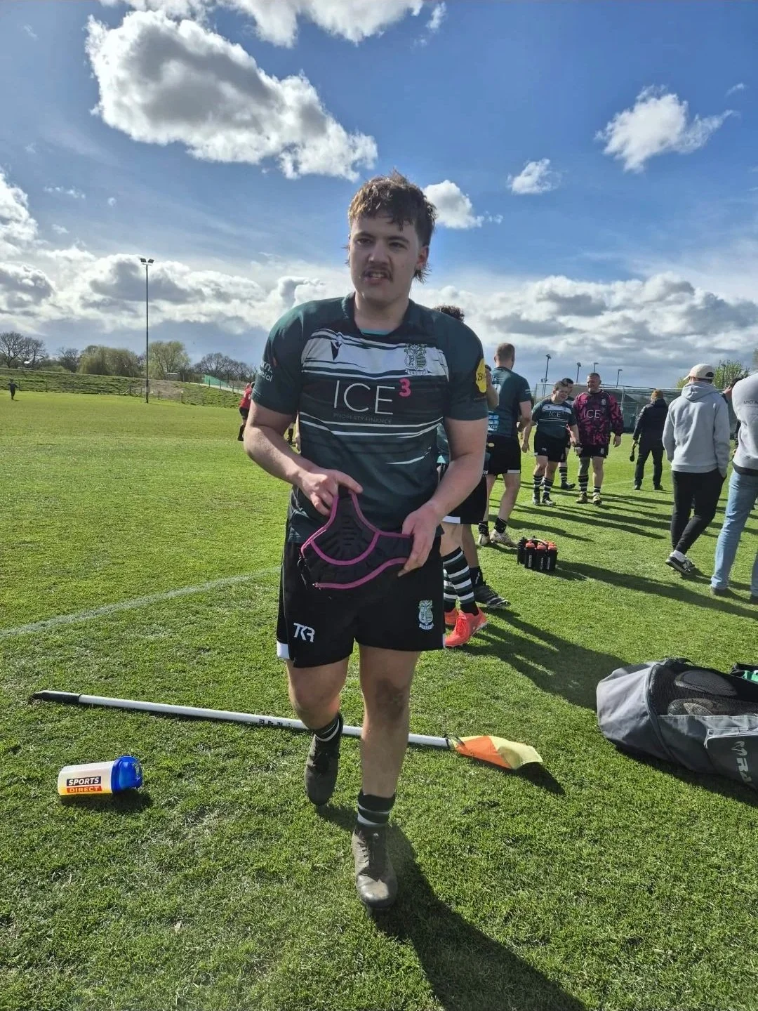 Rugby player in black uniform walking off the field holding a pink sports bra, with other players and coaches in the background on a sunny day.