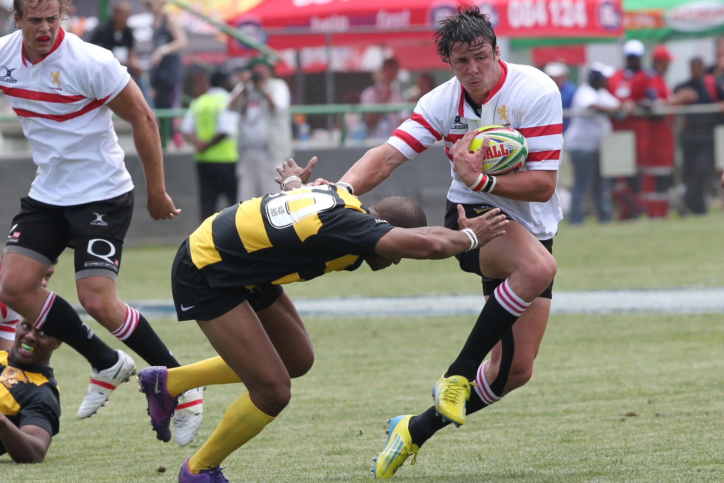A rugby match with players in action, featuring a player in a white jersey holding the ball while running, and other players tackling or trying to stop him on the field.