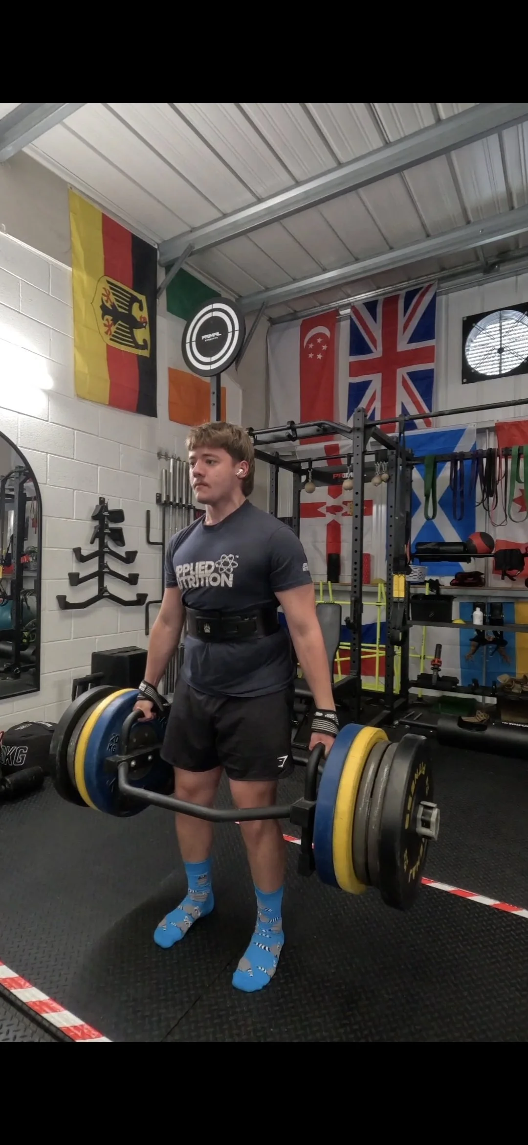 A man lifting a heavy barbell with weights in a gym, surrounded by flags including Germany, Ireland, Singapore, the UK, and others.