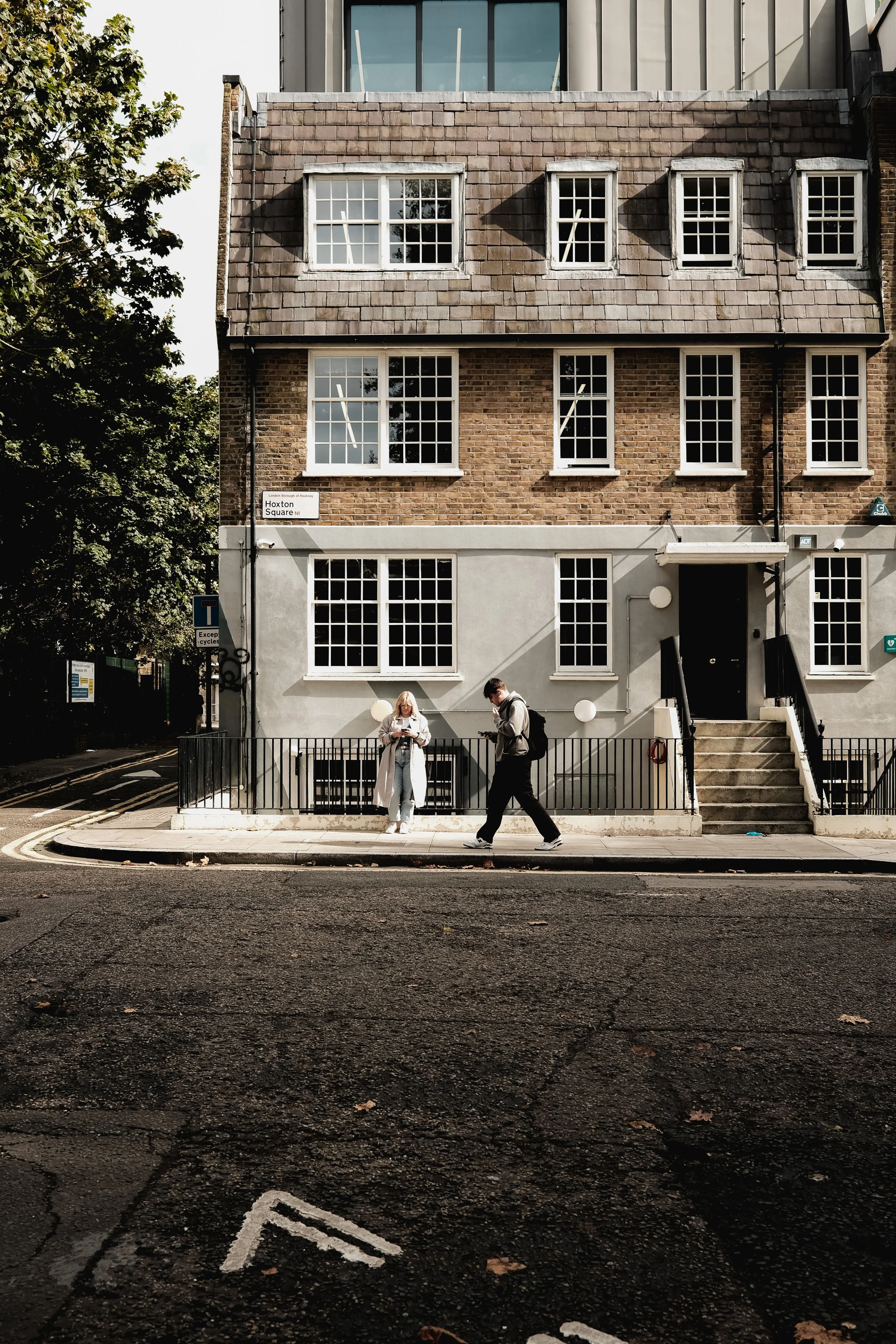 A three-story building with a brick facade and white-framed windows, with two people standing outside on the sidewalk near a metal barrier. One person, wearing a light-colored coat, is looking at their phone, while the other person, dressed in darker clothing, is walking past. There's a street sign that reads "Hoxton Square N1" on the building's corner, and a few trees visible on the left side of the image.