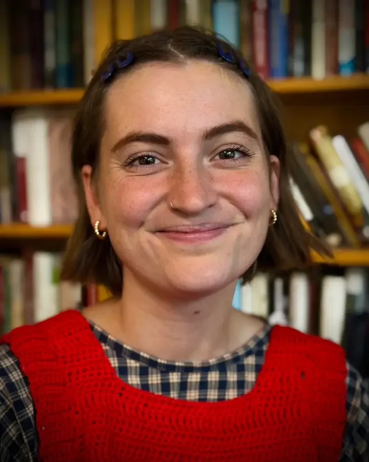 A young woman with short brown hair, wearing a red knit vest over a checkered shirt, smiling in front of a background of bookshelves in a library.