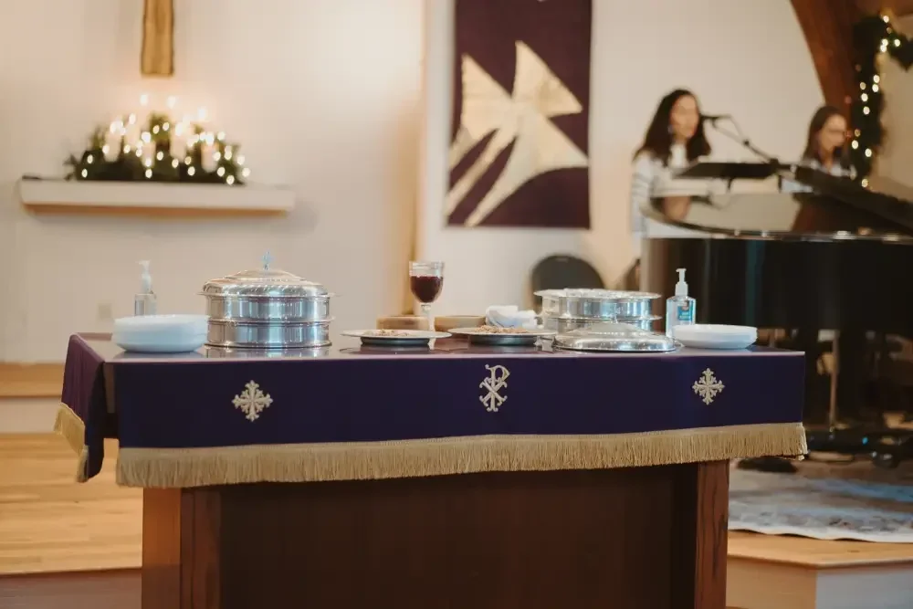 A table covered with a dark blue cloth with Christmas symbols, set for a religious or holiday event, with a glass of wine, stacked metal containers, and a bottle of hand sanitizer. In the background, two women are singing or speaking into microphones near a grand piano, with Christmas lights and decorations around them.