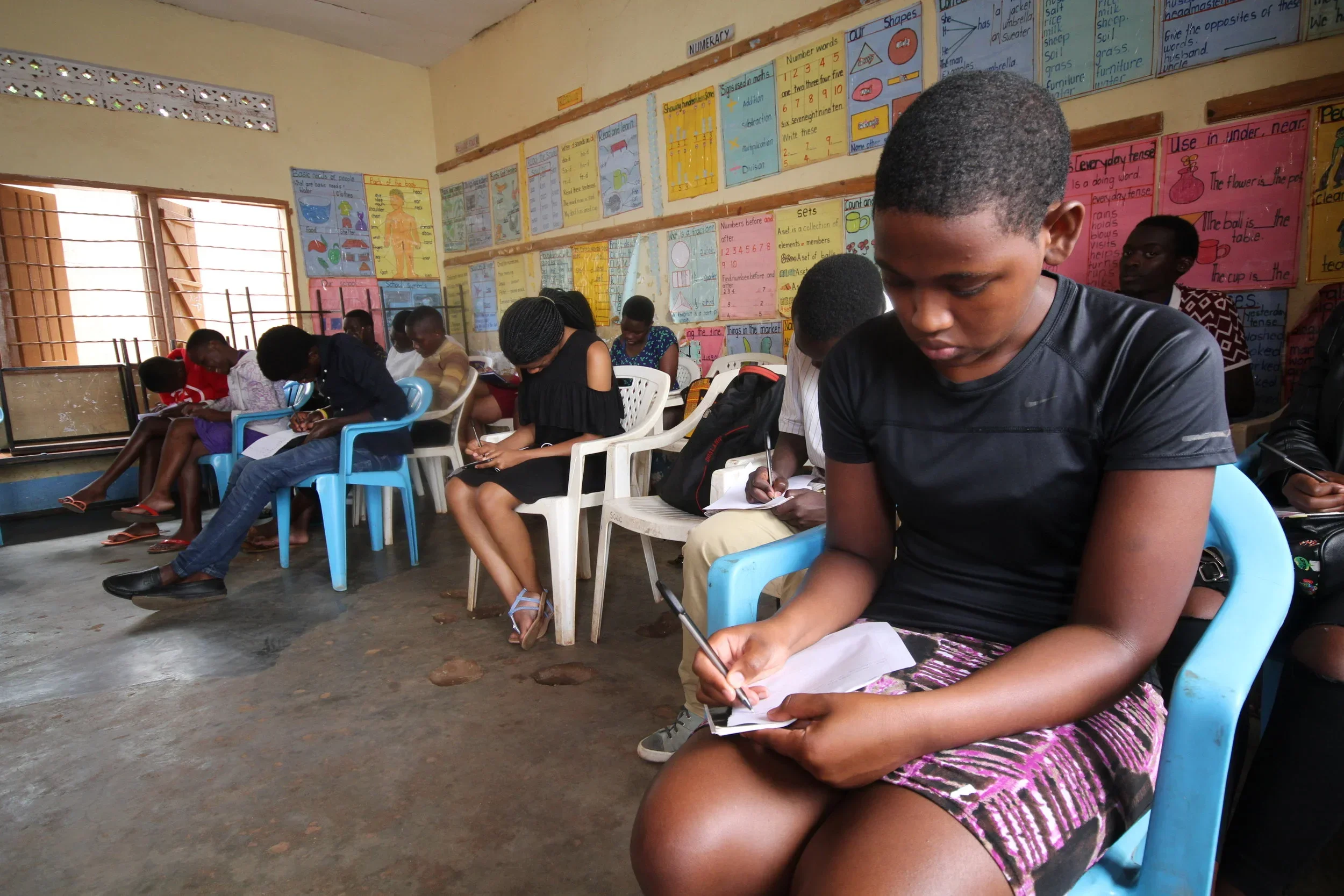 Students sitting on chairs and writing in notebooks in a classroom with educational posters on the wall.