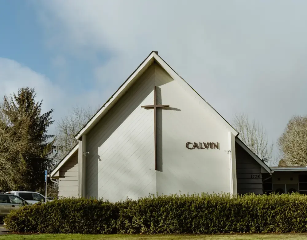 A church with a white exterior, a large cross on the front, and the name 'Calvin' on the wall. There are trees and parked cars nearby.