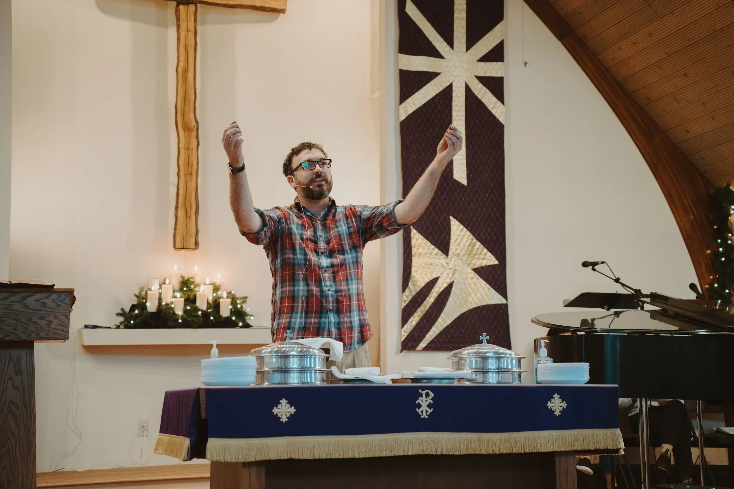 A man wearing glasses and a plaid shirt stands with his hands raised in a church decorated with a large American flag banner, a wooden cross, and candles.