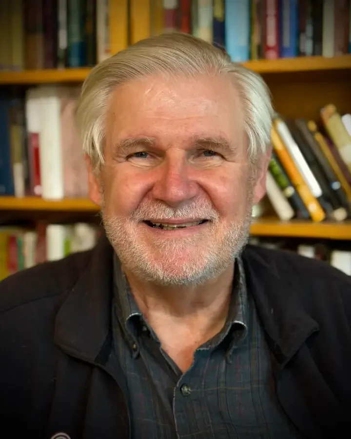 A smiling older man with a beard and gray hair, wearing a dark jacket and plaid shirt, standing in front of bookshelves filled with books.