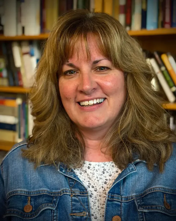 A smiling woman with shoulder-length, wavy, light brown hair posing in front of bookshelves in a library or bookstore, wearing a denim jacket over a white, patterned top.