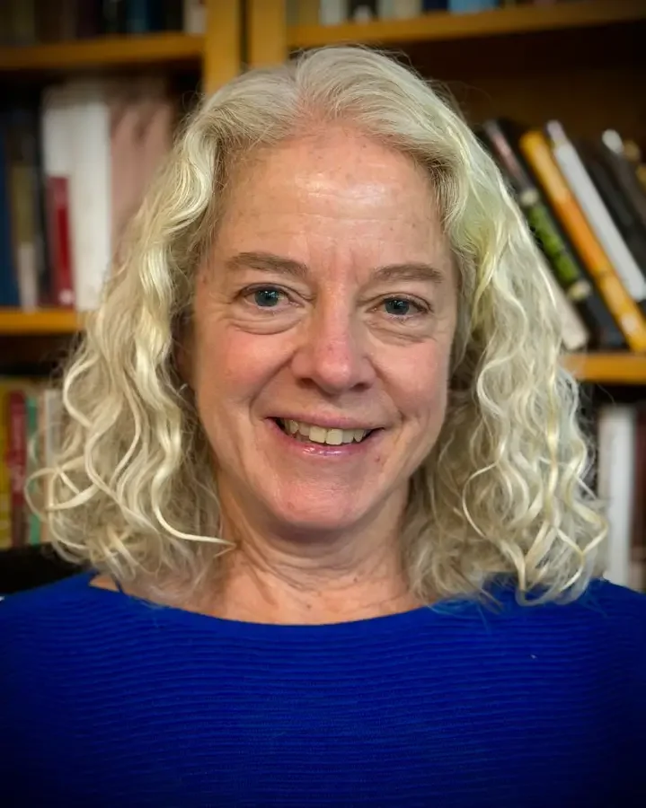 A smiling woman with curly, shoulder-length blonde hair, wearing a blue top, in front of a bookshelf filled with books.