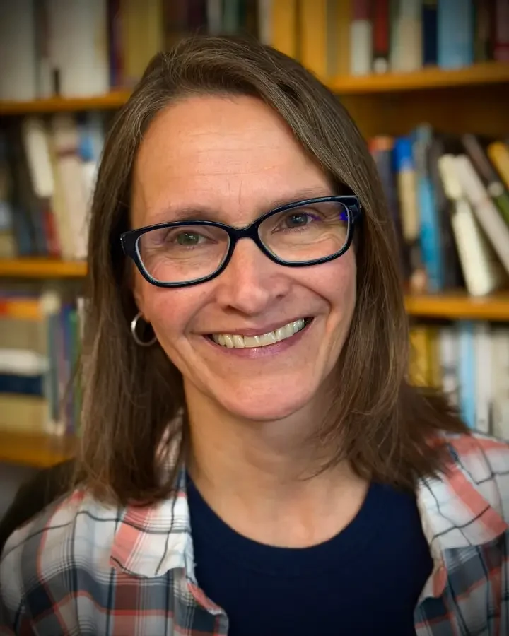 A smiling woman with shoulder-length brown hair, glasses, and hoop earrings, wearing a plaid shirt and standing in front of bookshelves in a library or bookstore.