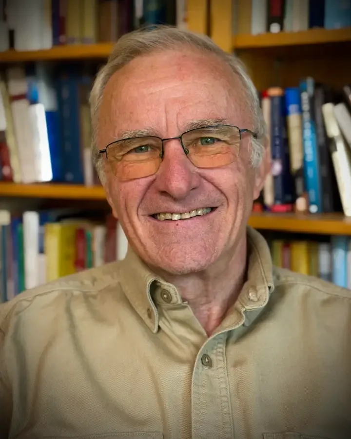 A smiling elderly man with gray hair, wearing glasses and a beige button-up shirt, standing in front of a bookshelf filled with books.