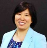 A woman with short black hair smiling, wearing a light blue blazer and a patterned blouse, against a dark background.