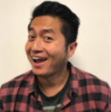A young man with short black hair smiling, wearing a red and black plaid shirt over a dark t-shirt.