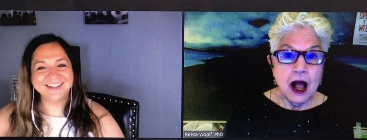 A screenshot of a virtual video call with two women. The woman on the left has long brown hair, is smiling, and wearing a sleeveless top. The woman on the right has short, curly blonde hair, glasses, and appears to be speaking with a surprised or expressive facial expression. The background of the woman on the right shows a painted landscape and her name tag reads "Reesa Woolf, PhD."