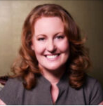 A woman with red hair and a friendly smile, wearing a blazer, in a professional setting.