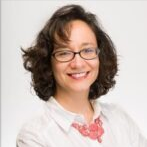 A smiling woman with curly dark hair and glasses, wearing a white shirt and a pink necklace.