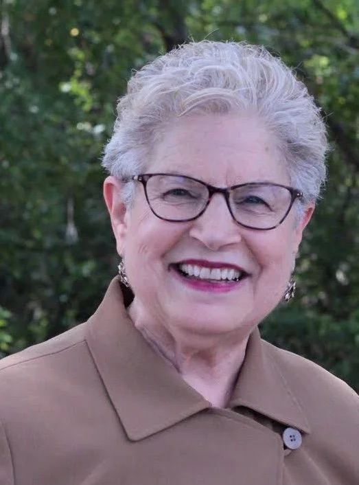 Smiling elderly woman with short curly gray hair, glasses, and earrings, wearing a tan coat, outdoors with green foliage in the background.