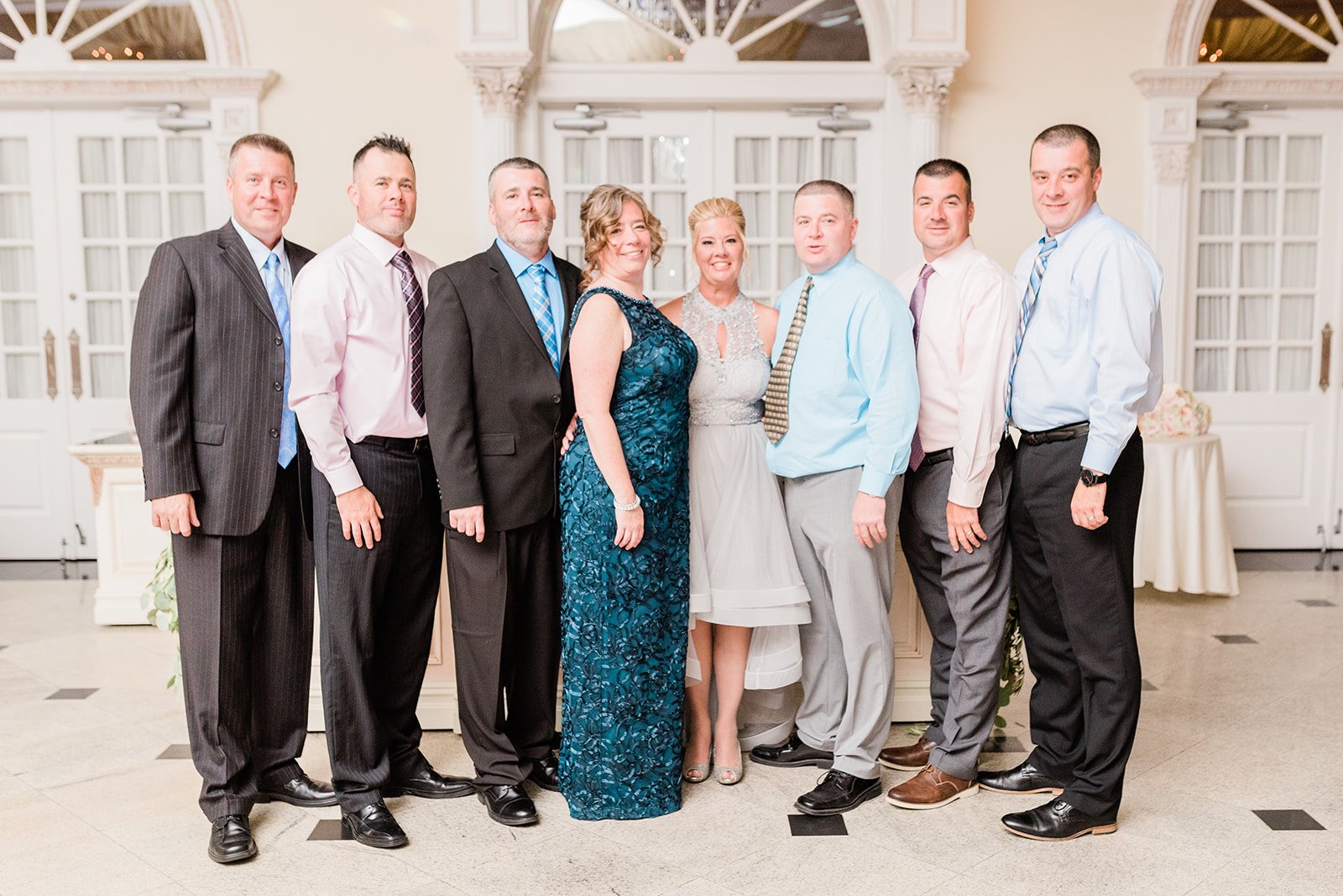 Group of nine people, six men and three women, dressed in formal attire, standing together indoors in front of windows and white decor, celebrating at an event or party.